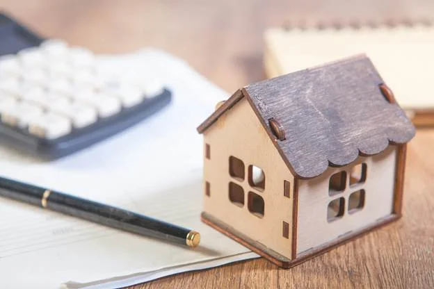 A small wooden house model, a pen, and a calculator rest on documents on a wooden desk, suggesting financial planning or property. | FintechZoom